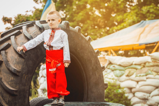 Ukrainian child boy in traditional national dress stands on roadblock near tire against background of sandbags. Concept of russian military invasion in Ukraine. War in Ukraine and Europe. Children and war. - Photo, Image