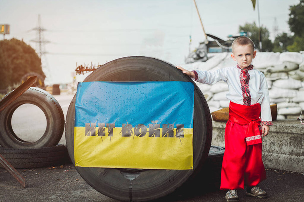 Ukrainian child boy in traditional national dress stands on roadblock near tire with inscription against background of sandbags. Concept of russian military invasion in Ukraine. War in Ukraine and Europe. Children and war. Translation of inscription: - Photo, Image