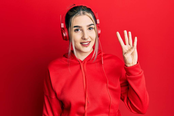Young modern girl wearing gym clothes and using headphones showing and pointing up with fingers number four while smiling confident and happy.  - Fotó, kép