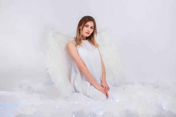 An attractive girl in a white tunic with large white wings behind her back poses while sitting in white luminous clouds. - Foto, Imagem
