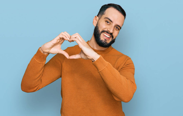 Молодий чоловік з бородою в повсякденному зимовому светрі посміхається, показуючи серцевий символ і форму руками. романтична концепція
.  - Фото, зображення