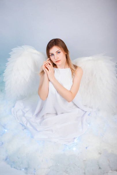 An attractive girl in a white tunic with large white wings behind her back poses while sitting in white luminous clouds. - Foto, imagen