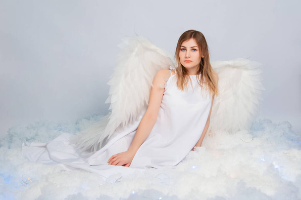 An attractive girl in a white tunic with large white wings behind her back poses while sitting in white luminous clouds. - Foto, afbeelding