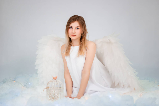 An attractive girl in a white tunic with large white wings behind her back poses while sitting in white luminous clouds. - Foto, Bild