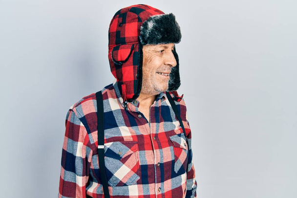 Bonito homem maduro usando chapéu de inverno com abas de orelha olhando para o lado com sorriso no rosto, expressão natural. rindo confiante.  - Foto, Imagem