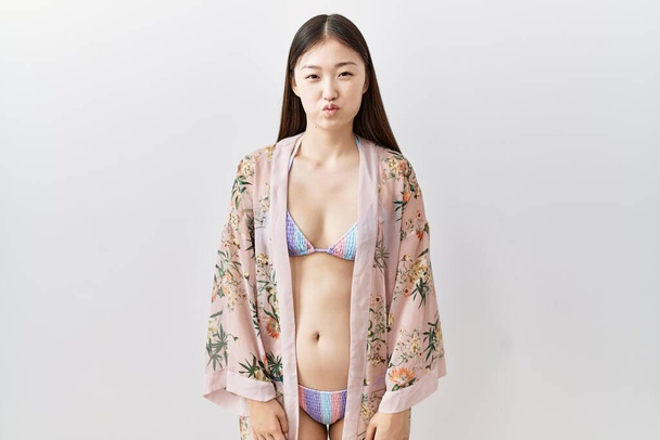 Młoda Azjatka w bikini z kwiatowym kimono nadętym policzkiem i śmieszną twarzą. usta napompowane powietrzem, szalony wyraz twarzy.  - Zdjęcie, obraz