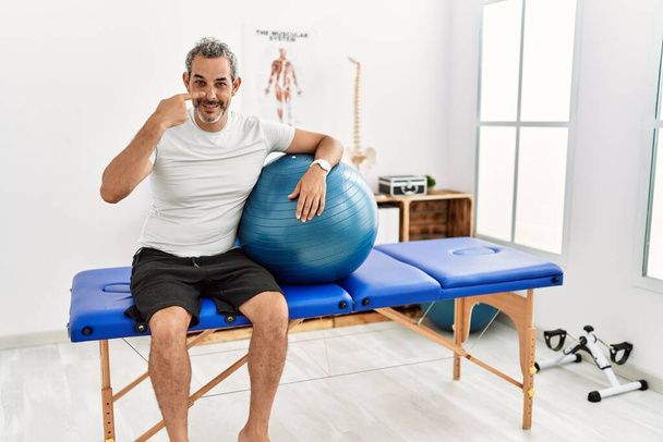 Hombre hispano de mediana edad en la clínica de recuperación del dolor sosteniendo la pelota de pilates señalando con el dedo a la mano y la nariz, sonriendo alegre. concepto de belleza  - Foto, Imagen