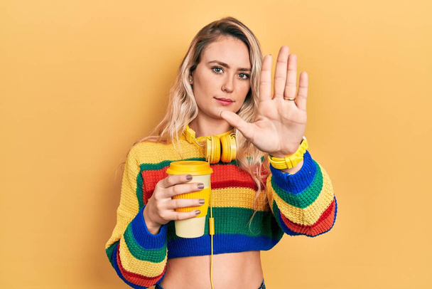 Mooie jonge blonde vrouw die een kop koffie drinkt met een koptelefoon die stopt met zingen met de palm van de hand. waarschuwingsuitdrukking met negatief en ernstig gebaar op het gezicht.  - Foto, afbeelding