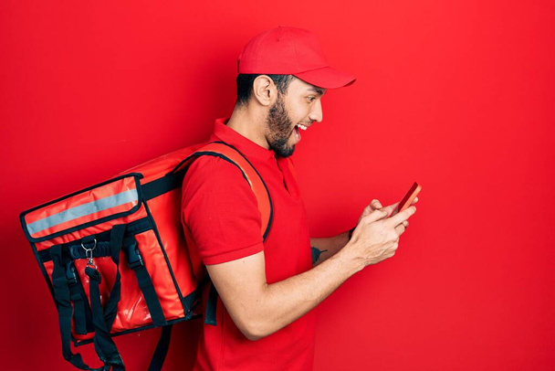 髭を生やしたヒスパニック系の男は、スマートフォンを使用して配達パッケージのバックパックを身に着けています。.  - 写真・画像