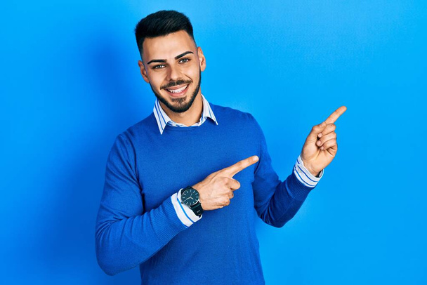 Joven hombre hispano con barba vistiendo suéter azul casual sonriendo y mirando a la cámara apuntando con dos manos y dedos hacia un lado.  - Foto, Imagen