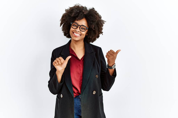 Αφροαμερικανή γυναίκα με αφρο μαλλιά που φοράει επαγγελματικό σακάκι και γυαλιά που δείχνουν προς τα πίσω με το χέρι και τους αντίχειρες προς τα πάνω, χαμογελώντας με αυτοπεποίθηση  - Φωτογραφία, εικόνα