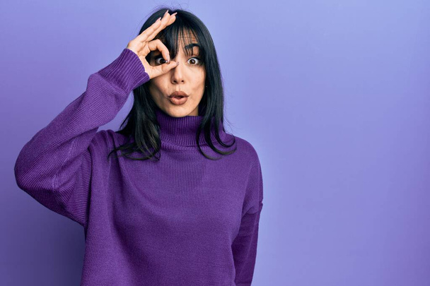 Young brunette woman with bangs wearing turtleneck sweater doing ok gesture shocked with surprised face, eye looking through fingers. unbelieving expression.  - Photo, Image