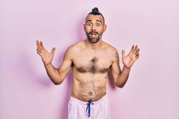 Joven hombre hispano sin camisa usando traje de baño celebrando loco y sorprendido por el éxito con los brazos levantados y los ojos abiertos gritando emocionado. concepto ganador  - Foto, imagen