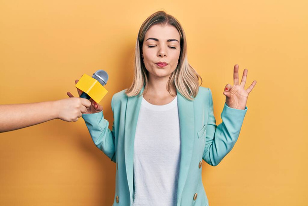 Beautiful caucasian woman being interviewed by reporter holding microphone relax and smiling with eyes closed doing meditation gesture with fingers. yoga concept.  - Photo, image