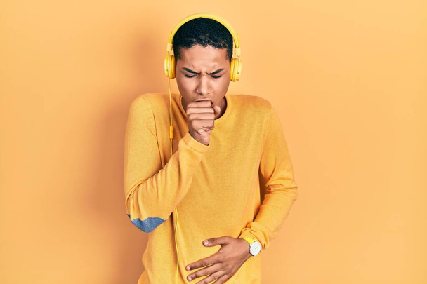 Νεαρός Αφροαμερικάνος που ακούει μουσική χρησιμοποιώντας ακουστικά που δεν αισθάνονται καλά και βήχει ως σύμπτωμα για το κρύο ή βρογχίτιδα. έννοια της υγειονομικής περίθαλψης.  - Φωτογραφία, εικόνα