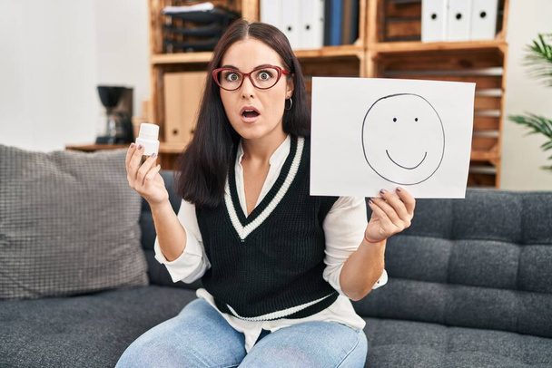 Young hispanic woman working on depression holding happy emotion on paper and pills afraid and shocked with surprise and amazed expression, fear and excited face.  - Photo, Image