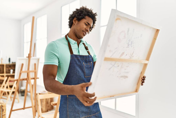 Νεαρός αφροαμερικανός καλλιτέχνης με σοβαρή έκφραση κρατώντας καμβά στο στούντιο τέχνης. - Φωτογραφία, εικόνα