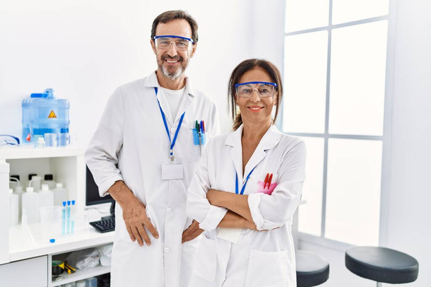 Mężczyzna i kobieta w średnim wieku partnerzy w mundurze naukowca stojący z bronią w ręku skrzyżowany gest w laboratorium - Zdjęcie, obraz