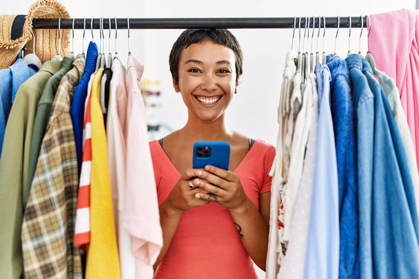 Νεαρή ισπανόφωνη γυναίκα με κοντά μαλλιά που ψάχνει ρούχα στη σχάρα ρούχων χρησιμοποιώντας το smartphone αναζητούν θετική και ευτυχισμένη στέκεται και χαμογελά με αυτοπεποίθηση χαμόγελο που δείχνει τα δόντια  - Φωτογραφία, εικόνα
