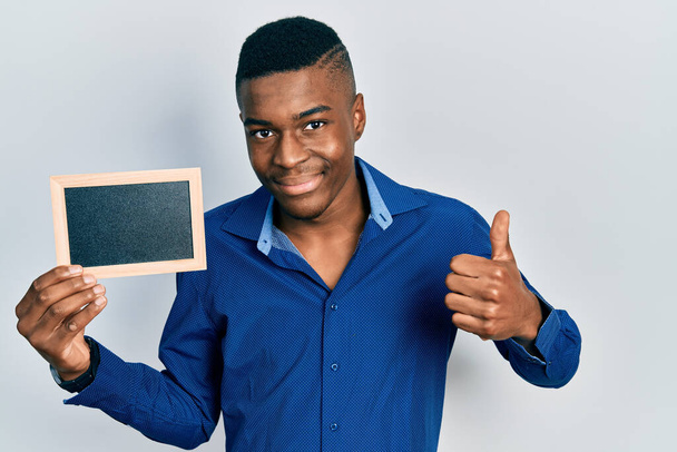 Молодой африканский американец держит пустую рамку улыбаясь счастливой и позитивной, большой палец вверх делает отлично и знак одобрения  - Фото, изображение