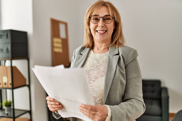 Middle age businesswoman smiling happy holding paperwork standing at the office. - Photo, Image