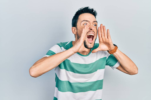 Joven hombre hispano usando ropa casual gritando enojado en voz alta con las manos sobre la boca  - Foto, Imagen