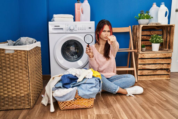 Young caucasian woman using magnifying glass glass looking for stains in dirty laundry pointing with finger to the camera and to you, confident gesture looking serious  - Photo, Image