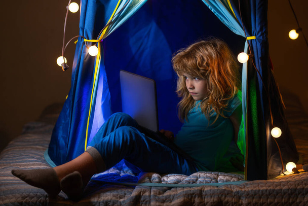 Little boy in tent bed looking at the laptop at night. Little hacker, young programmer. Child boy using computer at night in bed. - Фото, изображение