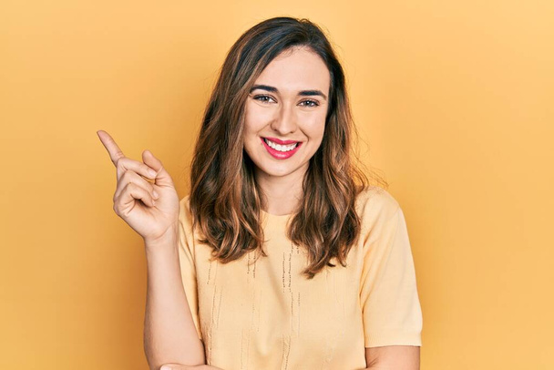 Jeune fille hispanique portant des vêtements décontractés souriant heureux pointant avec la main et le doigt sur le côté  - Photo, image