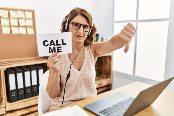 Middle age brunette woman wearing operator headset holding call me banner looking unhappy and angry showing rejection and negative with thumbs down gesture. bad expression.  - Foto, afbeelding
