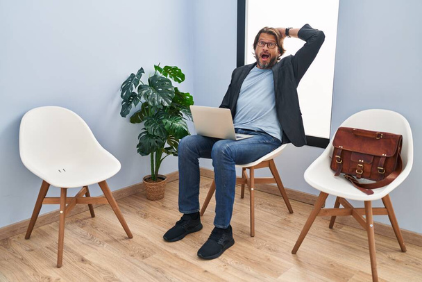 Όμορφος μεσήλικας που κάθεται στην αίθουσα αναμονής δουλεύοντας με το φορητό υπολογιστή τρελός και φοβισμένος με τα χέρια στο κεφάλι, φοβισμένος και έκπληκτος από το σοκ με ανοιχτό στόμα  - Φωτογραφία, εικόνα