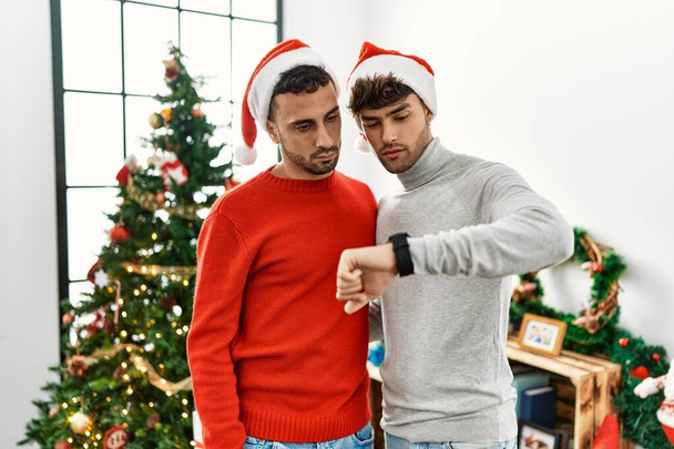 junges schwules Paar, das mit Hut am Weihnachtsbaum steht und die Uhrzeit auf der Armbanduhr kontrolliert, entspannt und selbstbewusst  - Foto, Bild