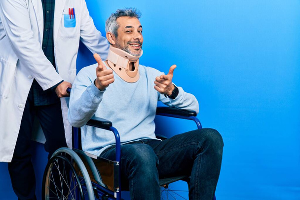 Όμορφος μεσήλικας άνδρας με γκρίζα μαλλιά σε αναπηρική καρέκλα φορώντας αυχενικό γιακά δείχνοντας τα δάχτυλα στην κάμερα με χαρούμενο και αστείο πρόσωπο. καλή ενέργεια και δονήσεις.  - Φωτογραφία, εικόνα