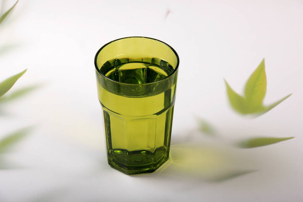 Zöld üveg vízzel és világos háttér növényi levelekkel. Top view zöld pohár víz napfény hosszú árnyék és fénytörési minták és pálmalevél trendi árnyék  - Fotó, kép