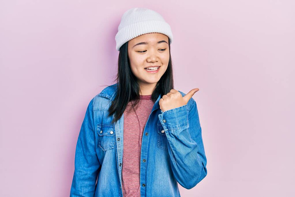 Fiatal kínai lány visel aranyos gyapjú sapka mosolyog boldog arc néz, és rámutatva oldalra hüvelykujjával felfelé.  - Fotó, kép