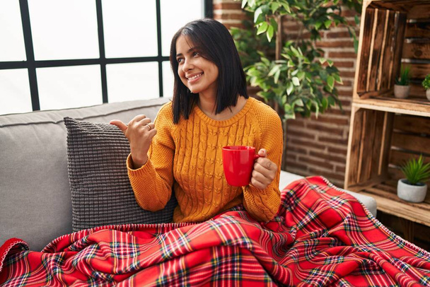 Genç İspanyol kadın kanepede oturmuş kahveyi yukardan içerken gülümsüyor ve başparmağıyla yan tarafı işaret ediyor..  - Fotoğraf, Görsel