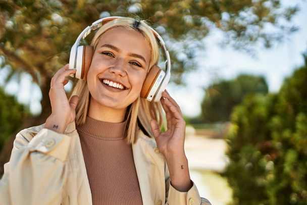 Νεαρή ξανθιά κοπέλα χαμογελά ευτυχισμένη με ακουστικά στην πόλη. - Φωτογραφία, εικόνα