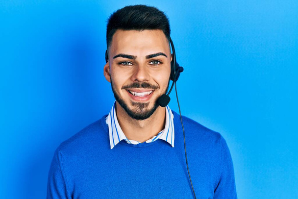 Fiatal spanyol férfi szakállas operátor headset-et visel a call center irodájában, pozitívan és boldogan áll és mosolyog magabiztos mosollyal a fogain.  - Fotó, kép
