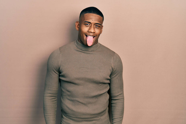 面白い表情で幸せに舌を突き出すカジュアルなタートルネックセーターを着た若い黒人男性。感情の概念.  - 写真・画像