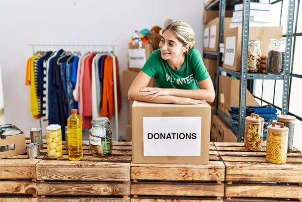 Νεαρή λευκή γυναίκα που φοράει στολή εθελοντή ακουμπισμένη στο κουτί δωρεών στο φιλανθρωπικό κέντρο - Φωτογραφία, εικόνα