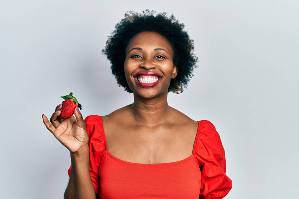 Joven mujer afroamericana sosteniendo fresa mirando positiva y feliz de pie y sonriendo con una sonrisa confiada mostrando los dientes  - Foto, Imagen