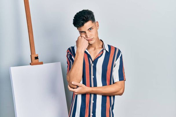 Молодий іспаномовний чоловік стоїть близько до порожнього полотна думаючи, що виглядає втомленим і нудним з проблемами депресії з схрещеними руками
.  - Фото, зображення