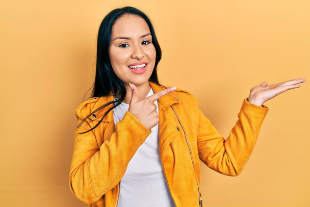 Beautiful hispanic woman with nose piercing wearing yellow leather jacket amazed and smiling to the camera while presenting with hand and pointing with finger.  - Photo, Image