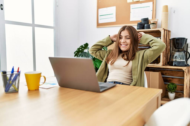 Молодая латиноамериканка улыбается уверенно расслабленной с руками на голове в офисе - Фото, изображение