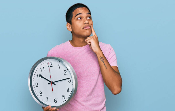 Young handsome hispanic man holding big clock serious face thinking about question with hand on chin, thoughtful about confusing idea  - Фото, изображение