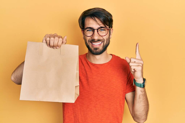 Jonge Latijns-Amerikaanse man met papieren zak met brood glimlachend met een idee of vraag wijzende vinger met gelukkig gezicht, nummer een  - Foto, afbeelding