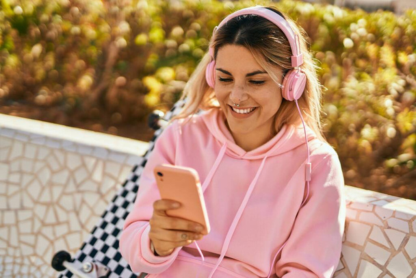 Joven chica rubia patinadora sonriendo feliz usando smartphone y auriculares en el parque. - Foto, imagen