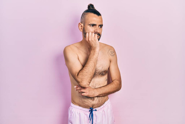 Junger hispanischer Mann ohne Hemd und Badeanzug, der gestresst und nervös aussieht, die Hände auf den Mund beißenden Nägeln. Angstproblem.  - Foto, Bild