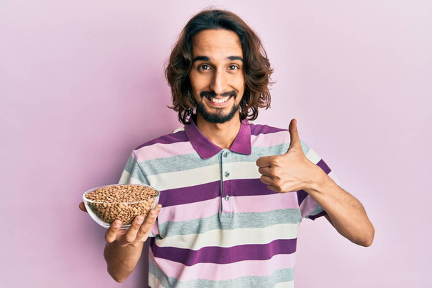 Young hispanic man holding lentils bowl smiling happy and positive, thumb up doing excellent and approval sign  - Foto, Bild
