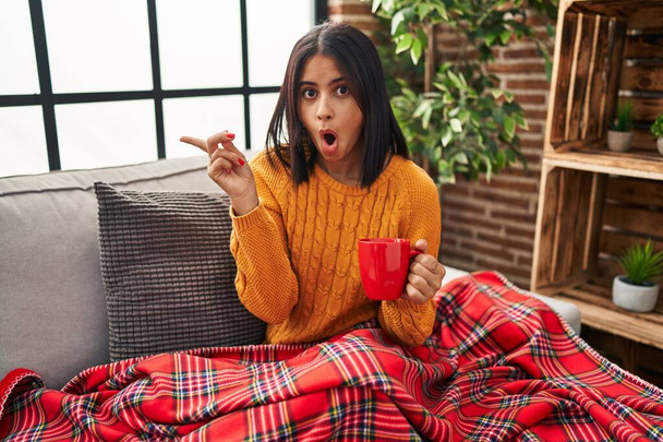 jonge Spaanse vrouw zit op de bank drinken van een koffie thuis verrast wijzend met de vinger naar de zijkant, open mond verbaasd uitdrukking.  - Foto, afbeelding